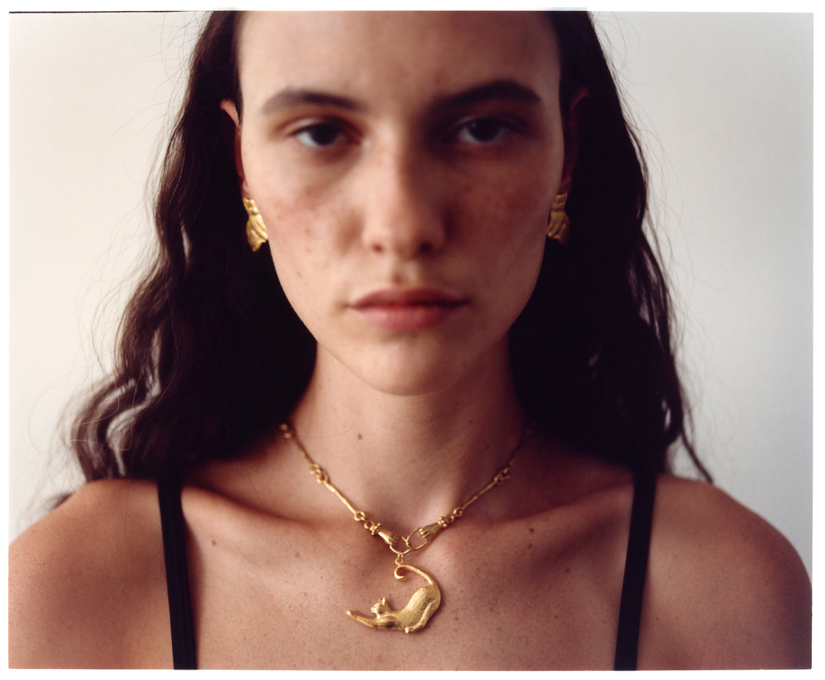 A close-up portrait of a model wearing an EFEK gold cat pendant necklace and gold earrings from the Cats of Istanbul collection.