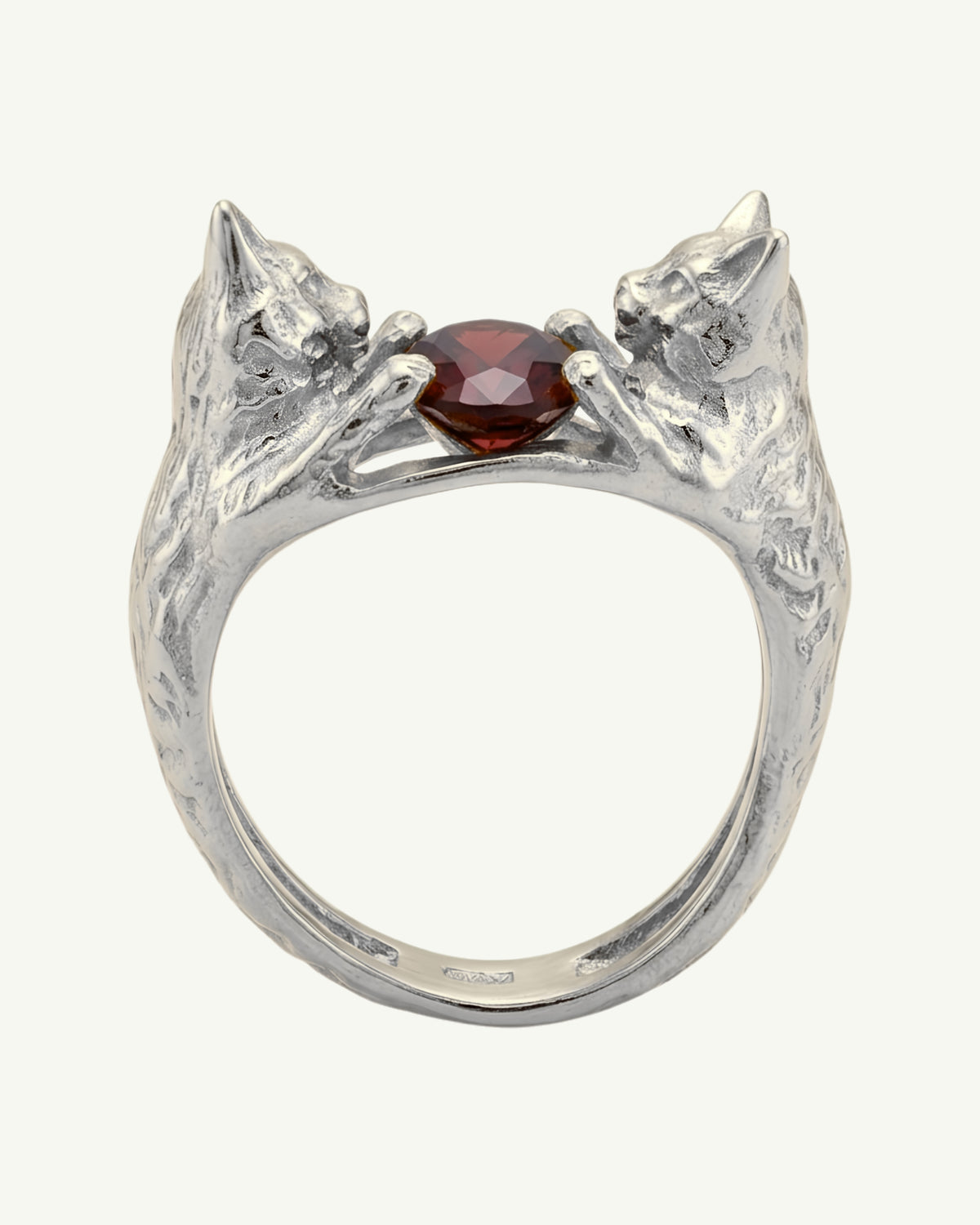 A silver ring featuring two sculpted cat heads facing each other around a central faceted garnet gemstone, shown on a plain light background.