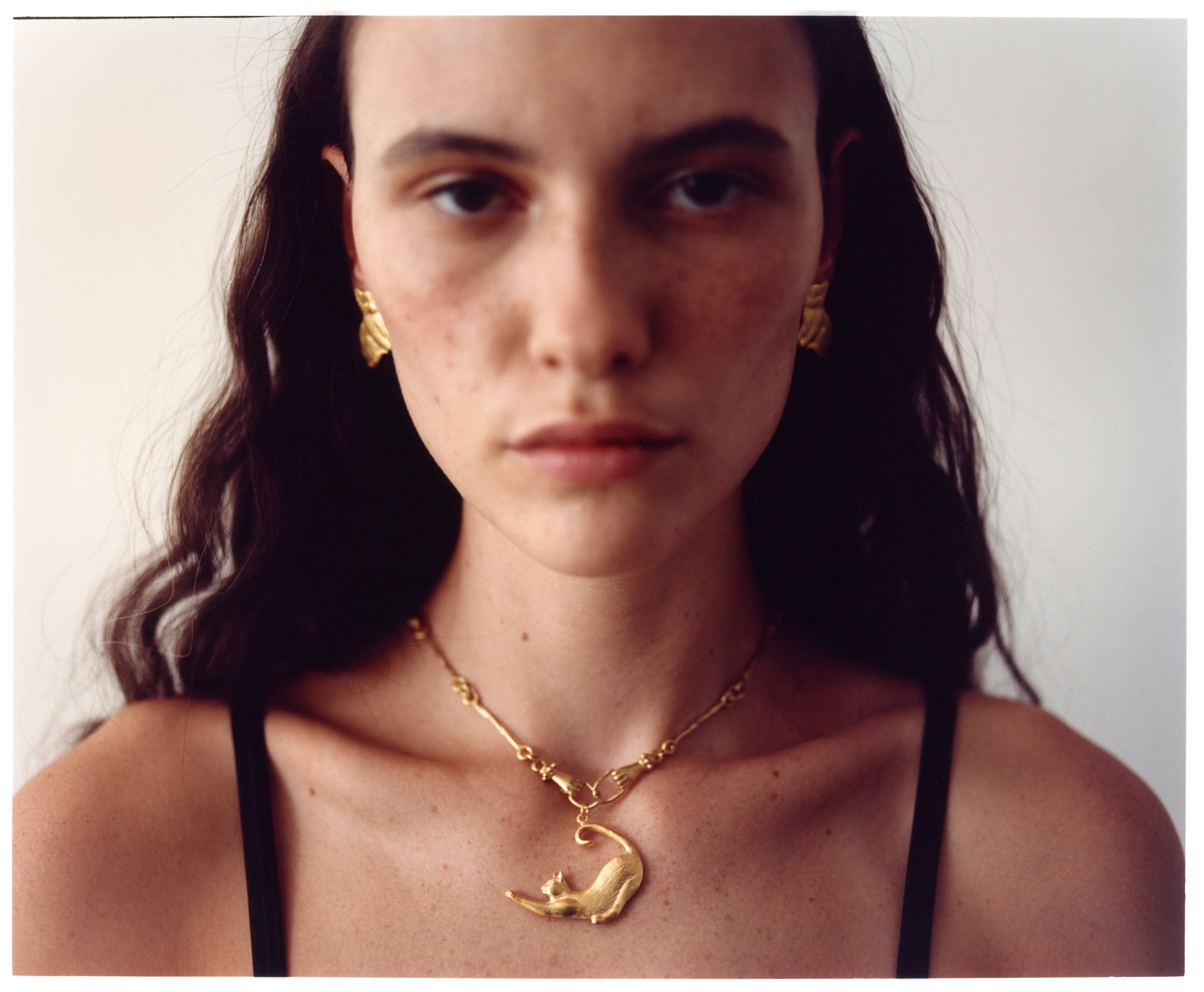 A close-up portrait of a model wearing an EFEK gold cat pendant necklace and gold earrings from the Cats of Istanbul collection.