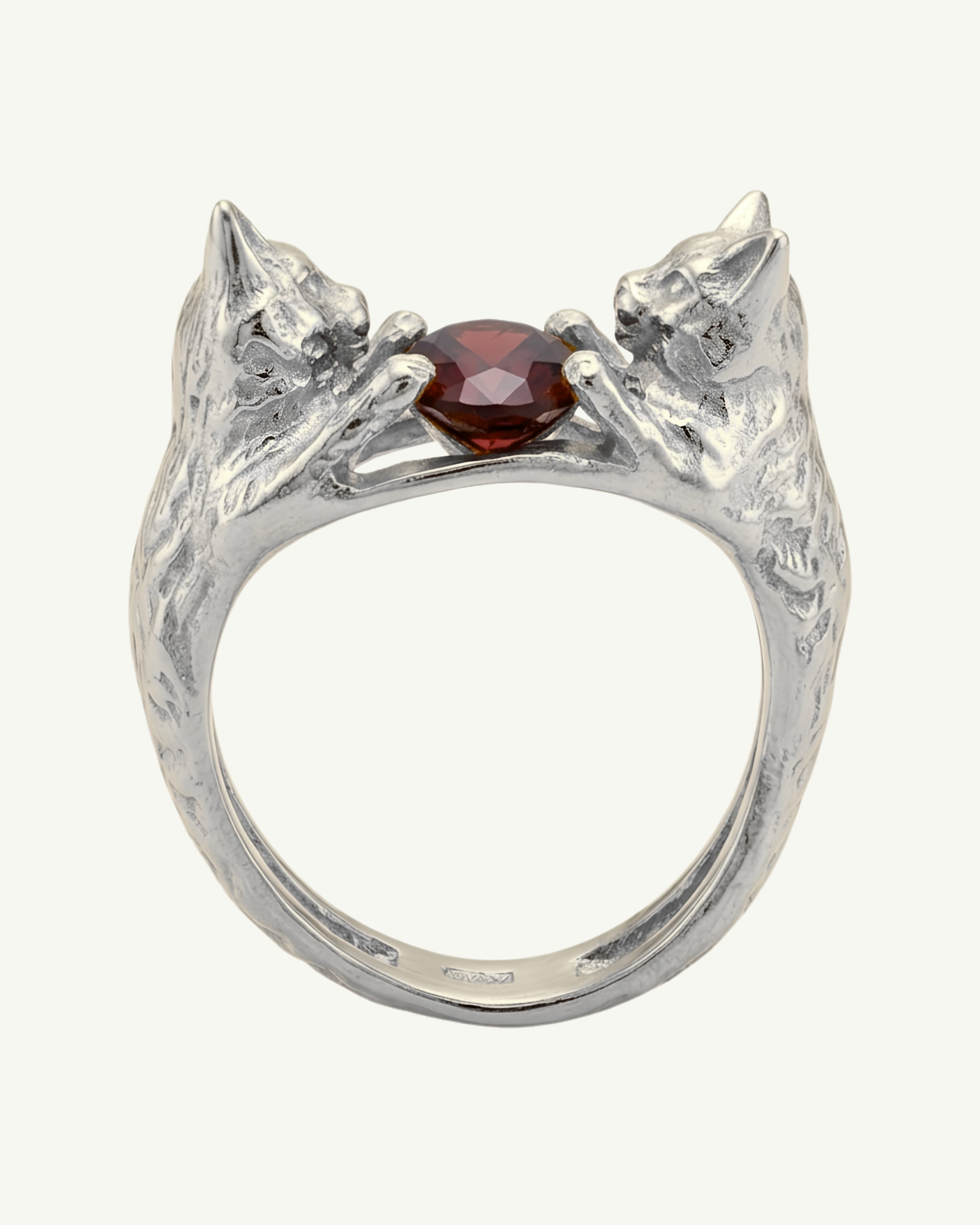 A silver ring featuring two sculpted cat heads facing each other around a central faceted garnet gemstone, shown on a plain light background.