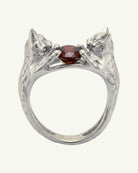 A silver ring featuring two sculpted cat heads facing each other around a central faceted garnet gemstone, shown on a plain light background.